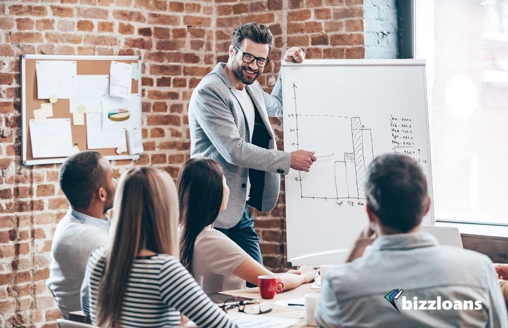 Business owner in glasses standing near whiteboard and discussing about their business growth strategy while his coworkers are listening and sitting at the table