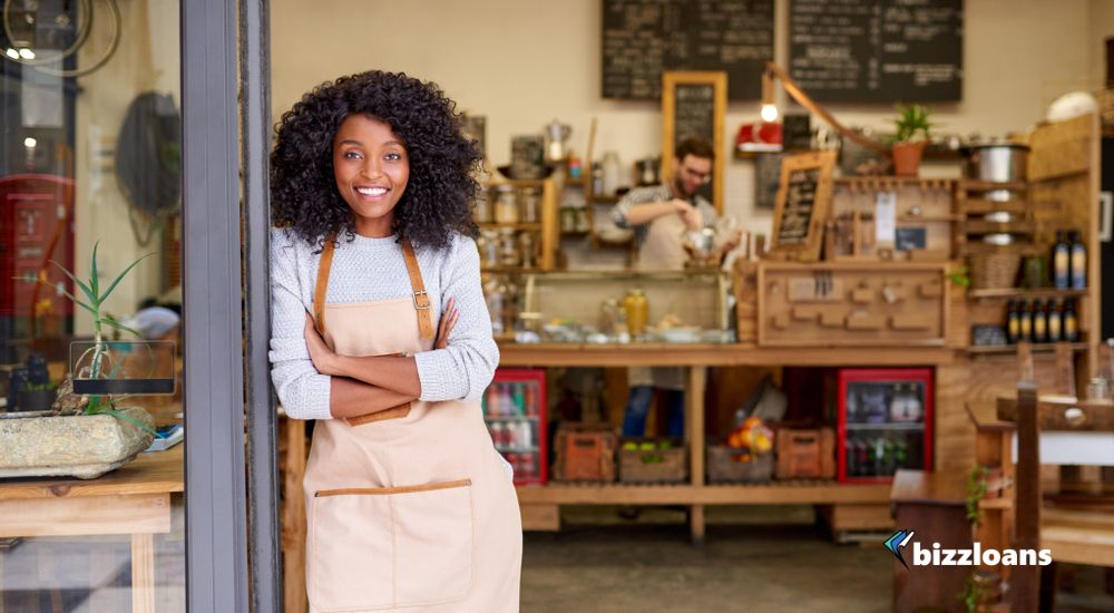 Portrait of a smiling young African American barista leaning with her arms crossed on the door of a trendy cafe