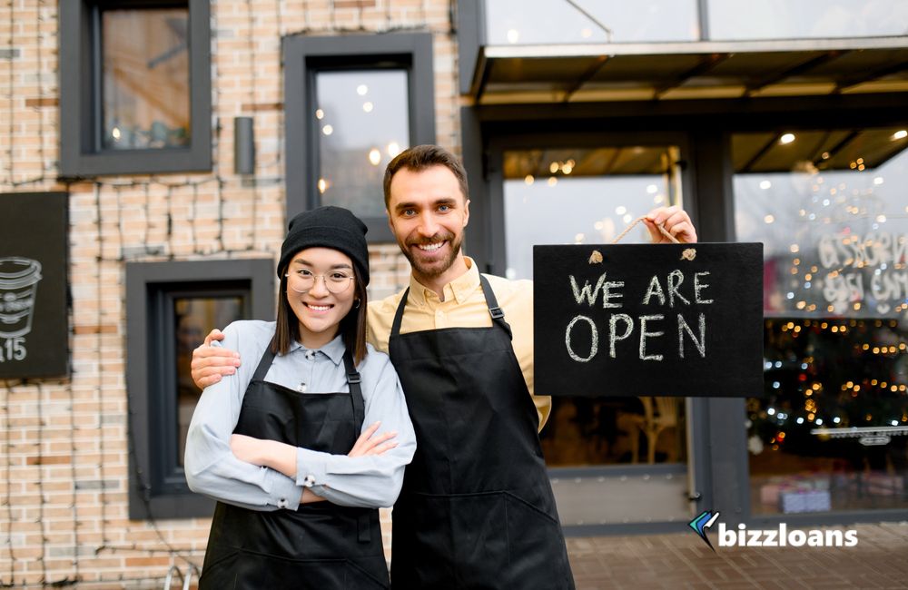 Two multiracial friendly waiters wearing uniform stand outside a restaurant, cafe or bar, showing signboard "We are OPEN", with pleasured smiling, welcome.